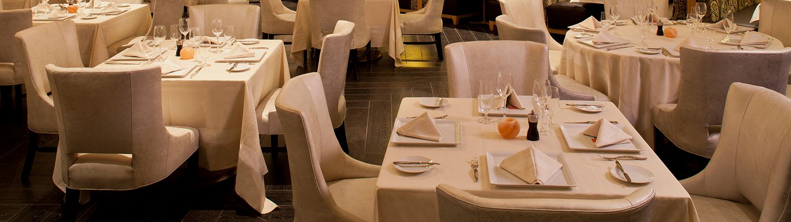 A dining room with several tables set with white tablecloths, chairs, wine glasses, napkins, and plates.