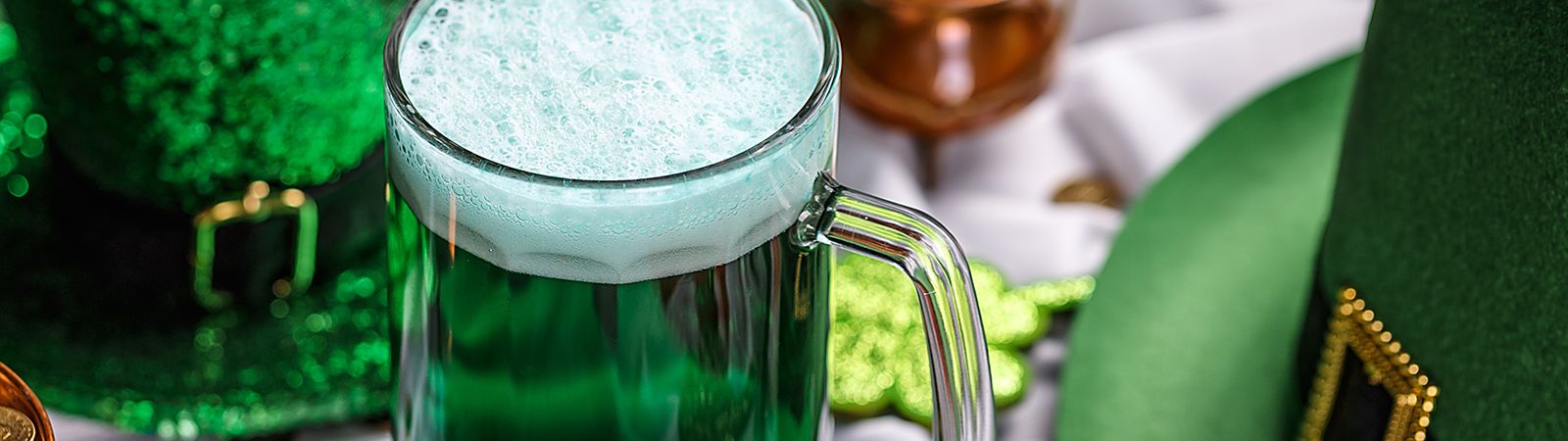 A glass mug of green beer with foam on top, surrounded by St. Patrick's Day decorations.